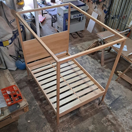 A 4-poster bed frame made of solid wood, constructed with a series of horizontal slats and a vertical support structure, displayed in a workshop setting with tools and materials around.