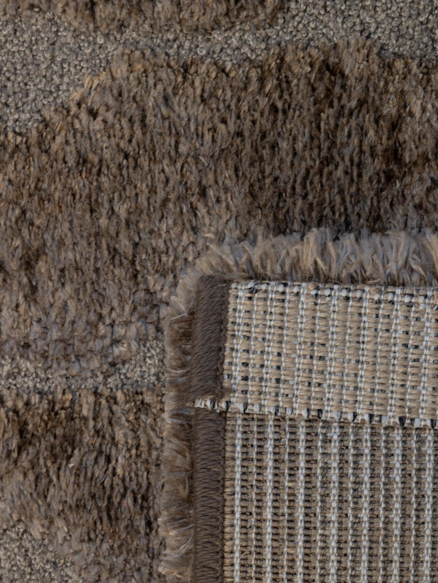 Close-up of a textured area rug showing a corner with a woven backing and fringe detail in taupe, brown, and beige tones.