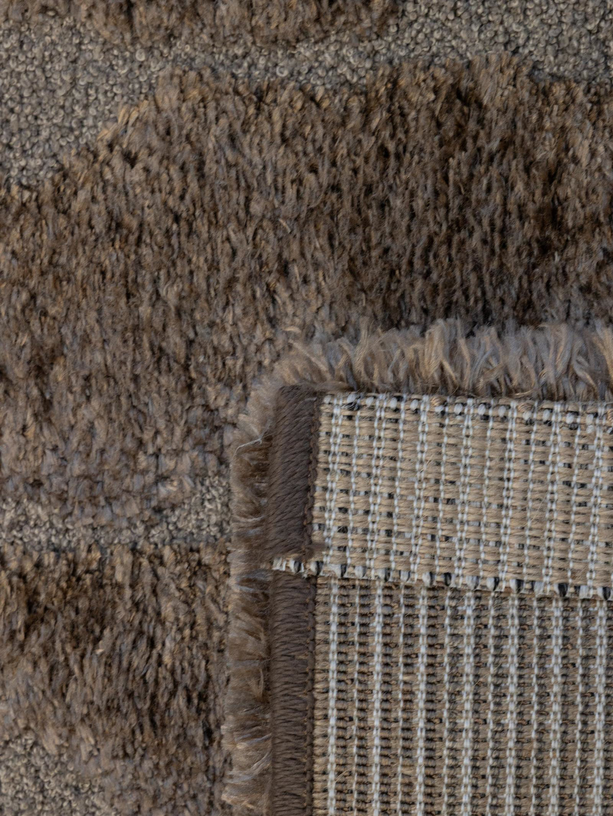 Close-up of a textured area rug showing a corner with a woven backing and fringe detail in taupe, brown, and beige tones.