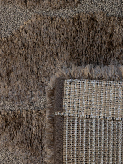 Close-up of a textured area rug showing a corner with a woven backing and fringe detail in taupe, brown, and beige tones.