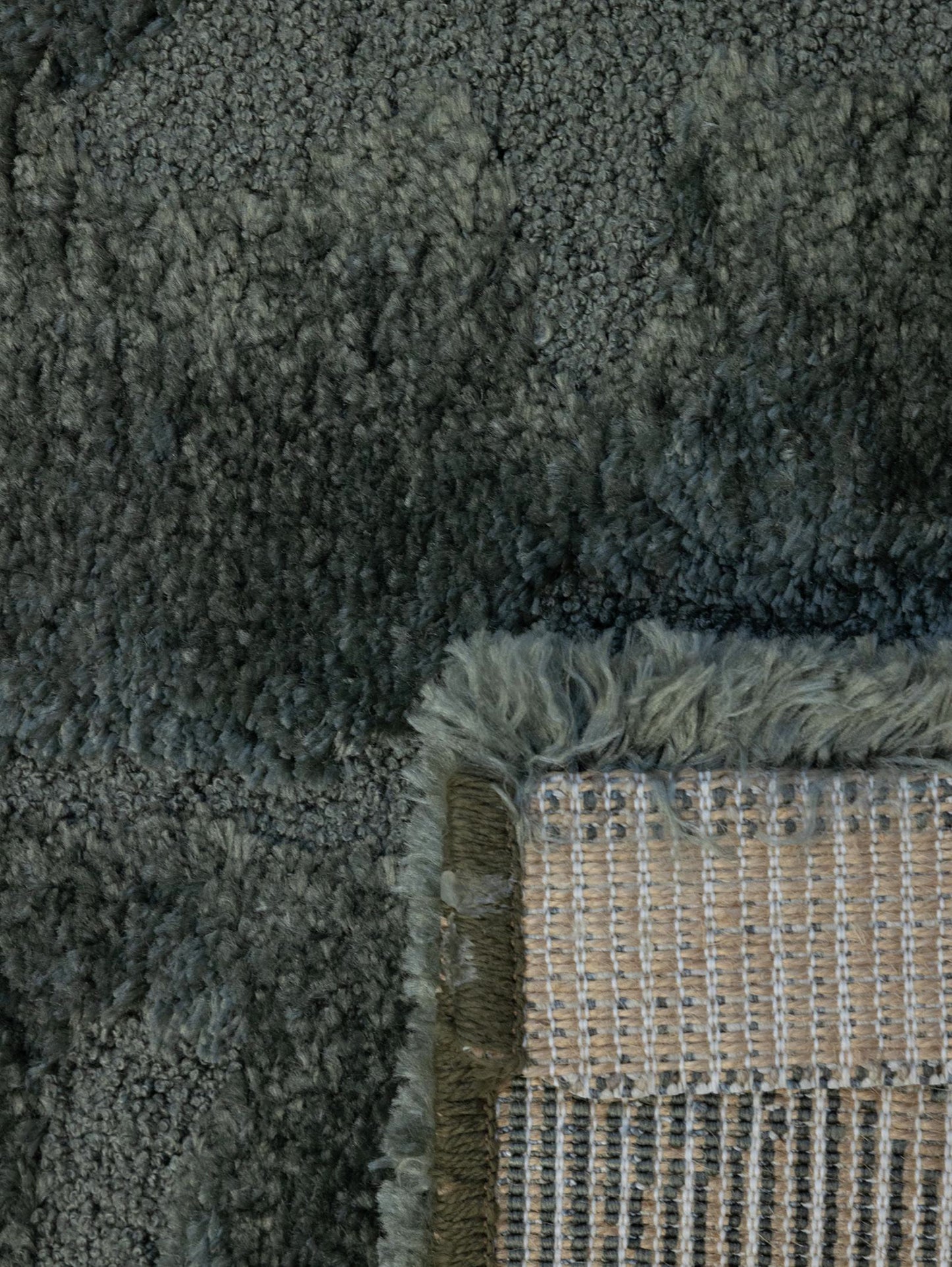 Close-up of a plush shag rug in a dark gray color, showing the thick pile texture and the corner where a beige woven backing and fringe are visible.