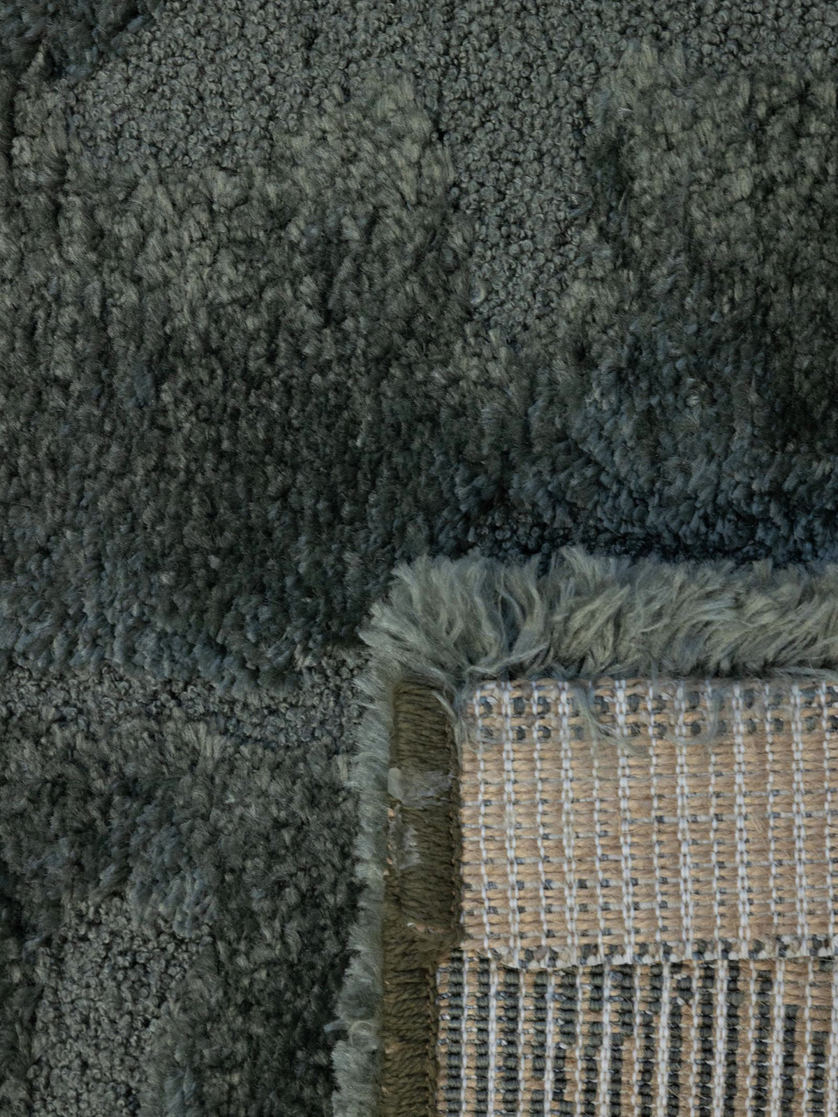 Close-up of a plush shag rug in a dark gray color, showing the thick pile texture and the corner where a beige woven backing and fringe are visible.