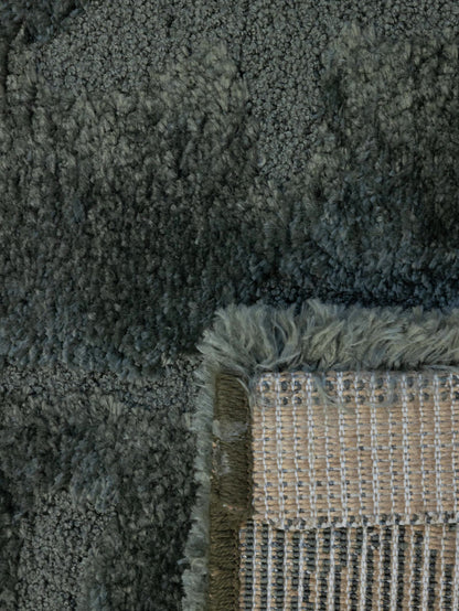 Close-up of a plush shag rug in a dark gray color, showing the thick pile texture and the corner where a beige woven backing and fringe are visible.